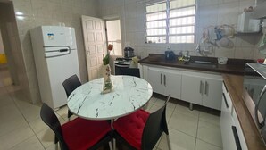 Una cucina con mobili bianchi, un frigorifero bianco, un tavolo da pranzo con sedie e una finestra con persiane.