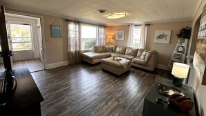 A living room with a sectional sofa, a coffee table, and a lamp. There is a door leading to a balcony, and a wall-mounted shelf with decorative items. - Bowline Knot - On the ICW - Dog Friendly! (Coinjock)