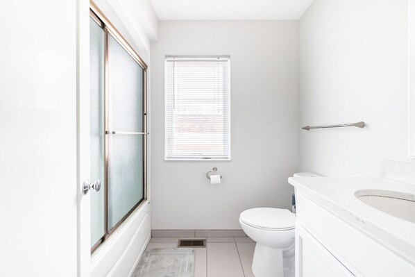 Un baño con inodoro, lavabo, ventana con persianas y ducha con puerta de cristal.