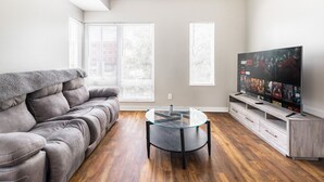 Een woonkamer met een grijze bank, een glazen salontafel, een televisie op een houten dressoir en grote ramen met zonwering.
