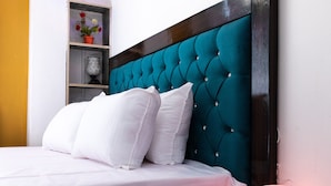 Una cama bien hecha con sábanas y almohadas blancas, un cabecero tapizado en color turquesa y una mesita de noche con una lámpara blanca.