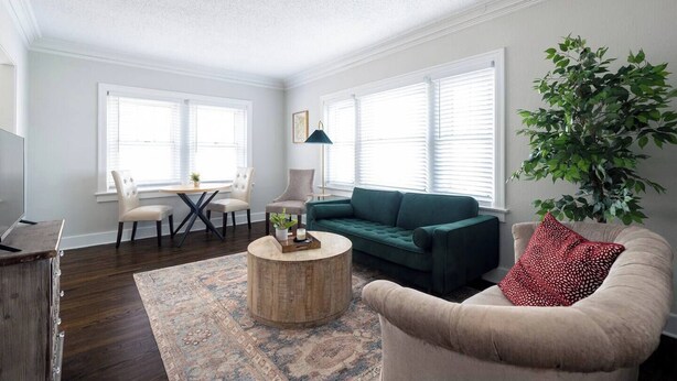 Ein Wohnzimmer mit einem grünen Sofa, einem beigefarbenen Sessel und einem runden Couchtisch. Es gibt einen Esstisch mit Stühlen, eine Lampe und eine große Pflanze.