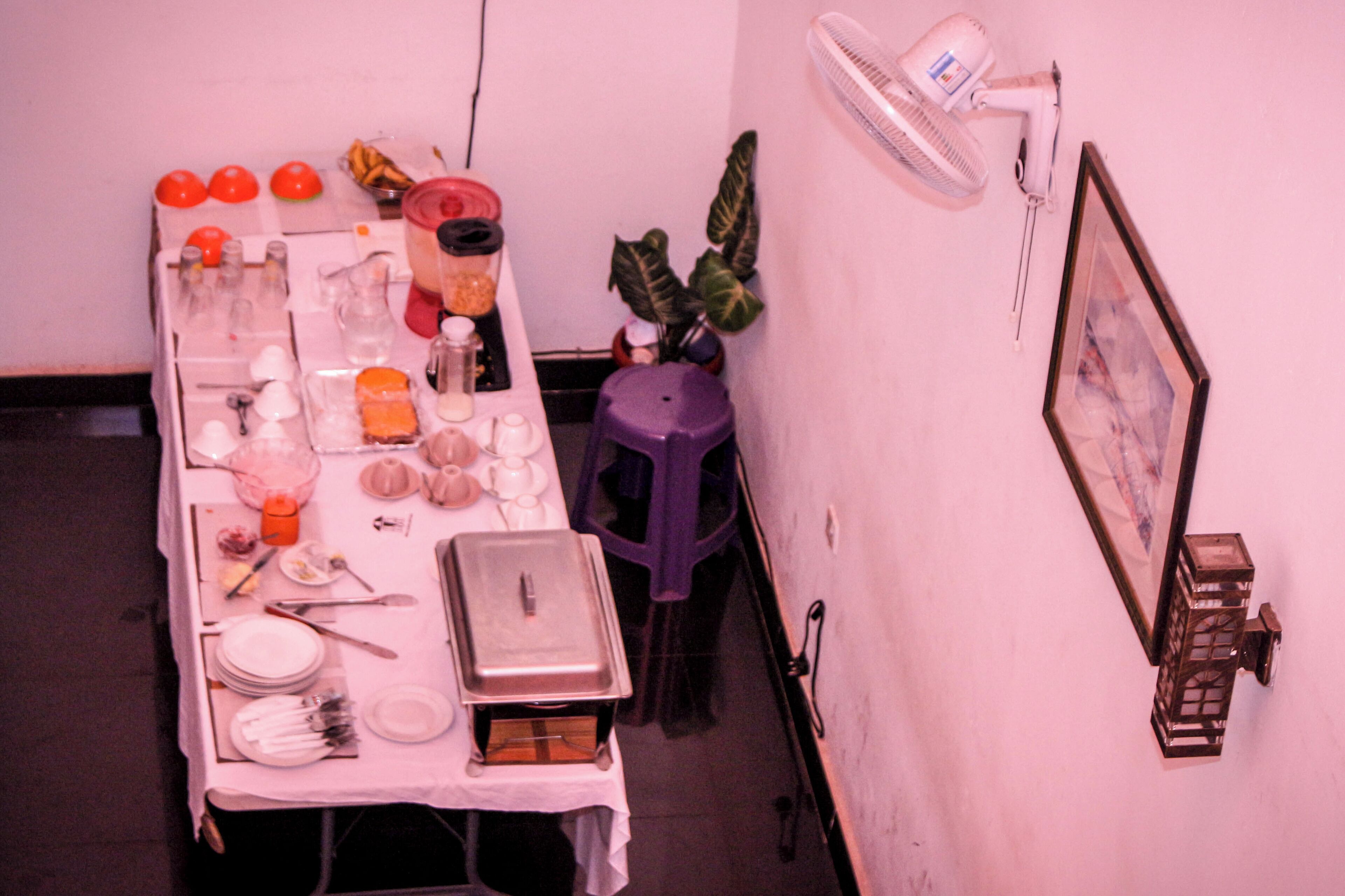 Une table à manger dressée avec des assiettes, des couverts et un récipient contenant de la nourriture, un ventilateur et un tableau encadré accroché au mur.