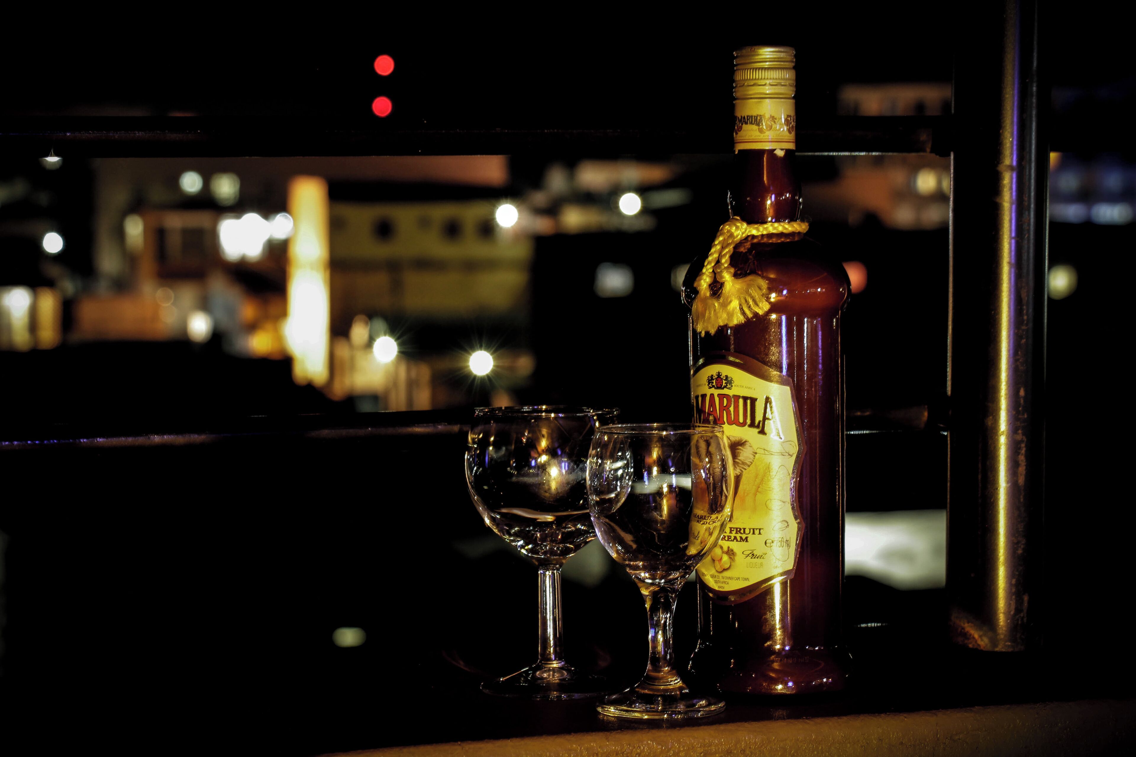 A bottle of liquor and two glasses on a bar counter with a cityscape view in the background.