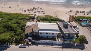 Un edificio costero con techo plano, junto a una playa con sombrillas y gente.