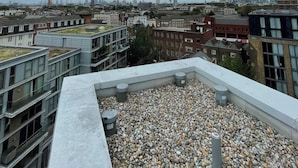 En takterrass med ett grusunderlag, omgiven av moderna byggnader och en stadsbild i bakgrunden.