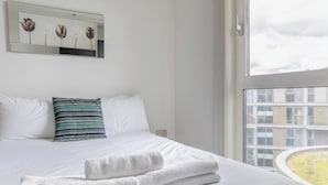 Ett hotellrum med en stor säng, vita sängkläder, en randig kudde och utsikt över byggnader genom ett fönster.