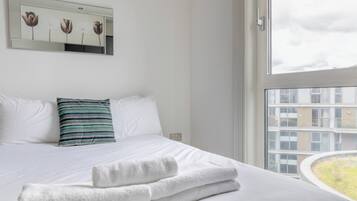 A hotel room with a large bed, white bedding, a striped pillow, and a view of buildings through a window.