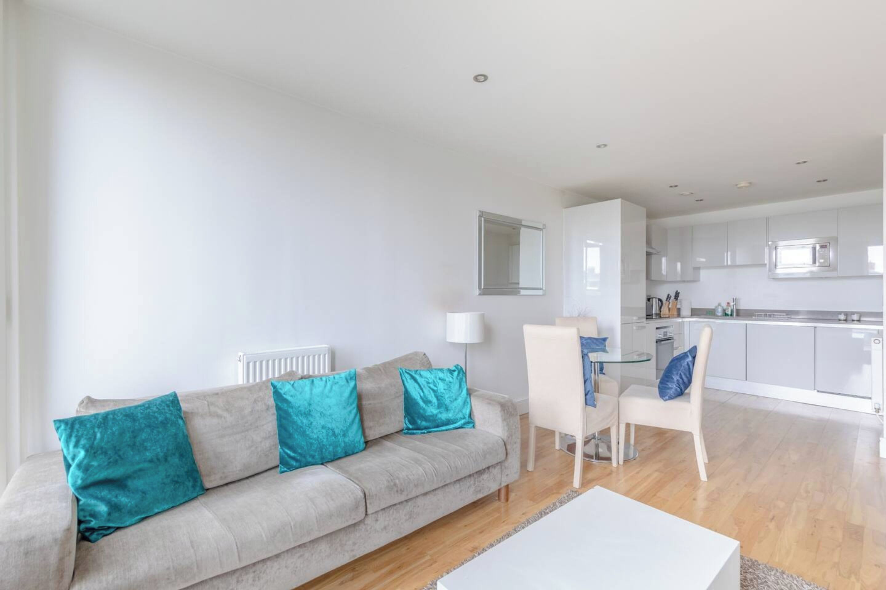 A modern living room with a grey sofa, teal cushions, a white coffee table, and a dining area with a white table and chairs.