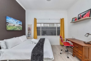 A hotel room with a bed, a desk, a chair, a window with yellow curtains, and a picture on the wall.