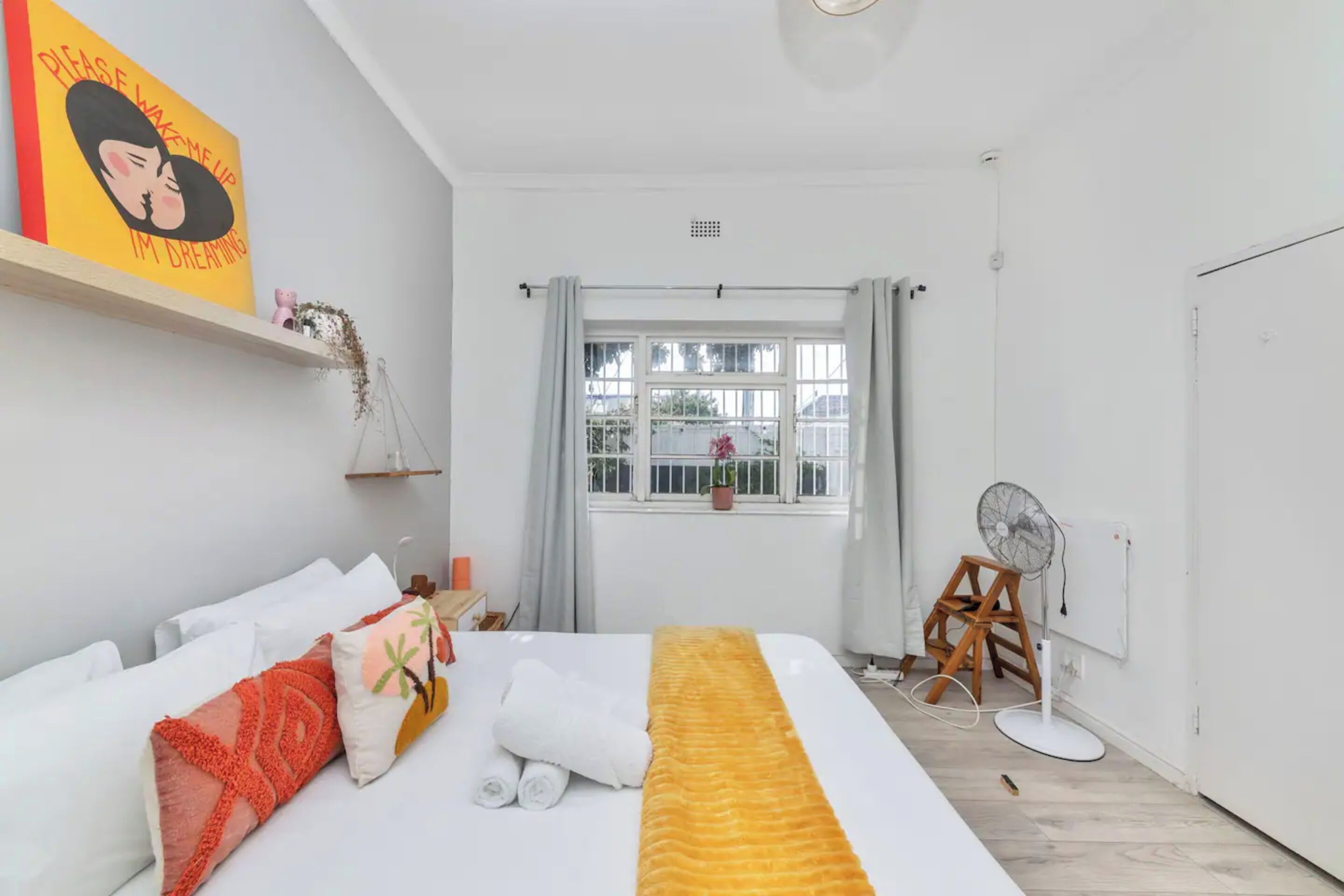 A hotel room with a bed, a window with curtains, a fan, and a wall-mounted shelf with a picture.