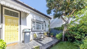Une maison avec une porte jaune, un petit patio avec une table et des chaises, et un jardin agrémenté d’un arbre et de plantes en pot.
