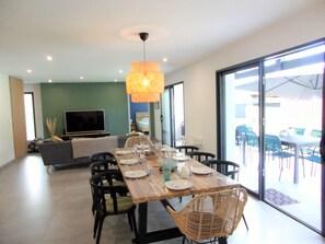 Un comedor con una mesa de madera dispuesta para cuatro personas, rodeada de sillas de mimbre, un televisor y un sofá al fondo.