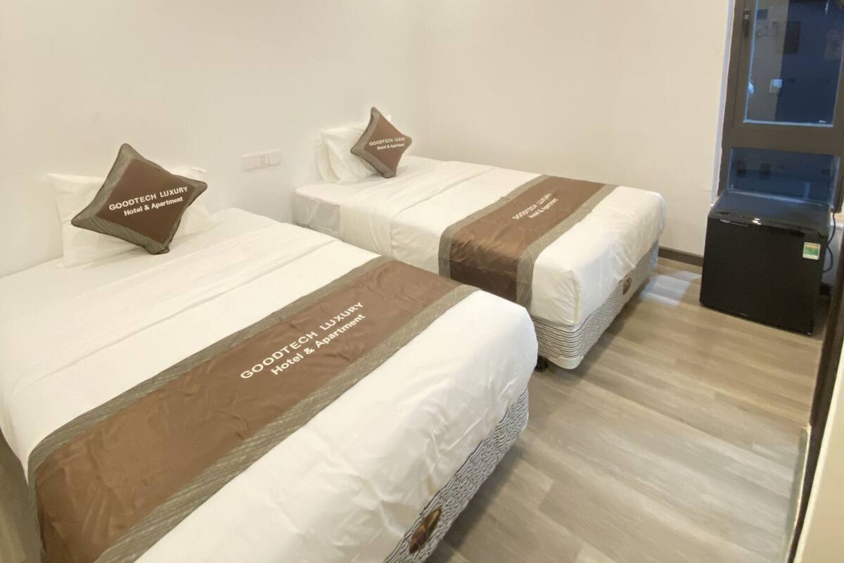 Zwei Betten in einem Hotelzimmer mit Holzboden und weißen Wänden. Jedes Bett ist mit einem Kopfkissen und einer Decke mit dem Namen des Hotels ausgestattet.
