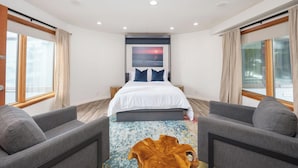 Una habitación de hotel moderna con una cama grande, dos sillones, una mesa de centro de madera y un cuadro de una puesta de sol sobre el océano.