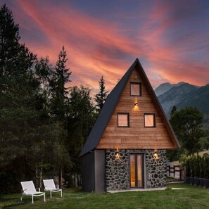 A modern, triangular wooden house with stone accents, surrounded by a lush green landscape and mountains in the background. - Chalet Tir Longe (Piemonte)