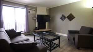 Ein Wohnzimmer mit Sofa, Sessel, Glastisch und einem Flachbildfernseher auf einem Standfuß.