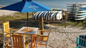 Ein Sitzbereich direkt am Strand mit Tisch und Stühlen, einem blauen Sonnenschirm und am Strand liegenden Kajaks.