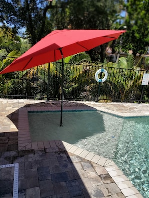A swimming pool with a red umbrella, surrounded by a brick border and a metal fence with a lifebuoy.