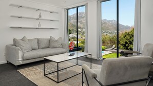 Ein stilvolles Wohnzimmer mit Sofa, Couchtisch und Sessel. Weiße Regale sind an der Wand angebracht, ein großes Fenster bietet Blick auf Berge und ein kleiner Tisch mit einem dekorativen Gegenstand.