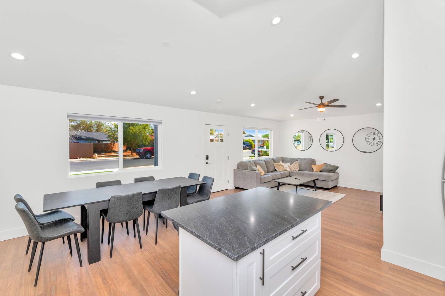 Une cuisine moderne avec un îlot central, un coin repas avec une longue table et des chaises, un canapé gris et un ventilateur de plafond.
