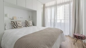 A modern bedroom with a large bed, a built-in wardrobe, and a window with sheer curtains.