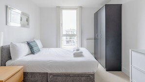 A modern bedroom with a bed, bedside table, wardrobe, and a window with a view. - Stylish One BDR Flat Close to Greenwich (London)