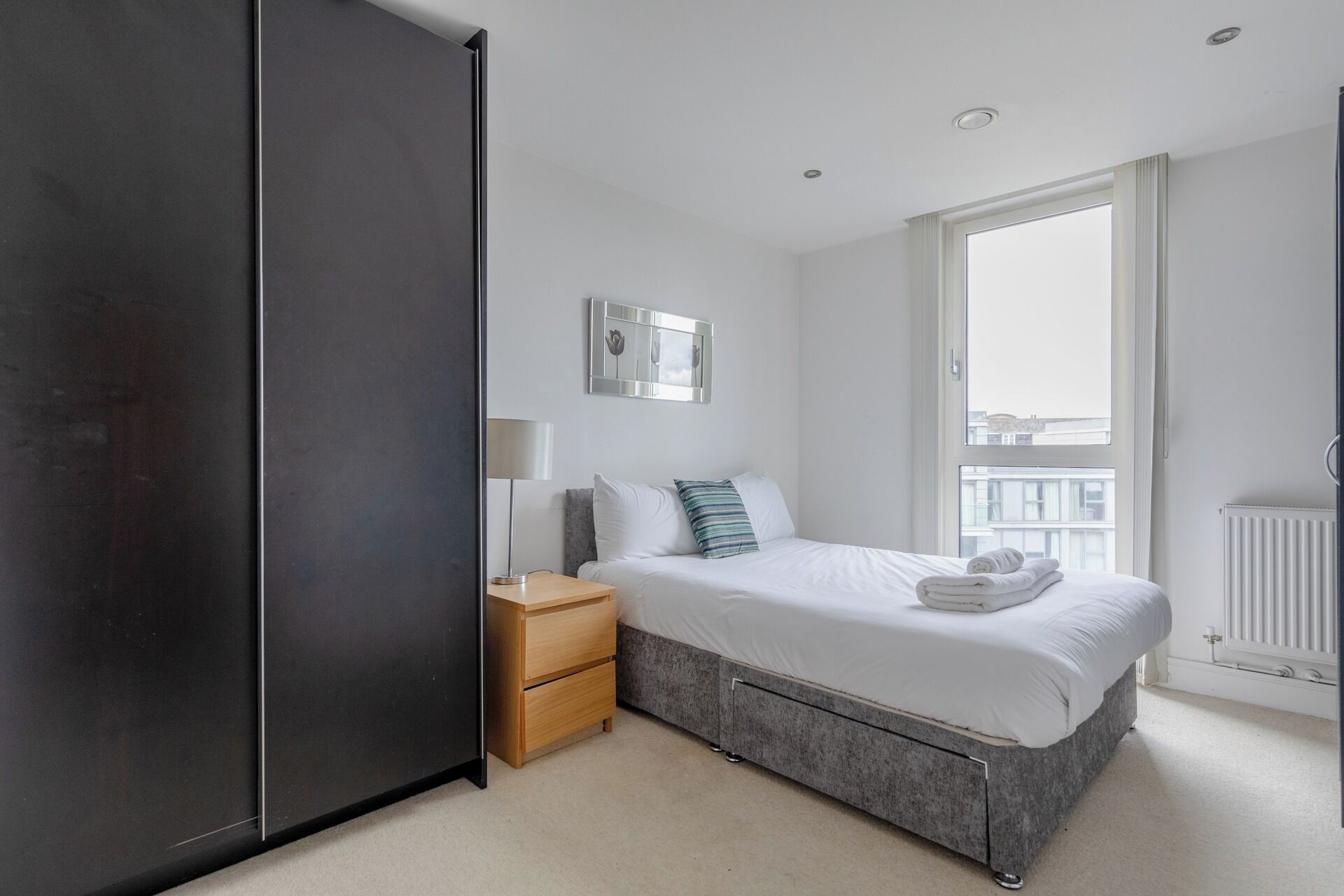 Quarto moderno com cama, mesa de cabeceira, abajur e uma janela ampla.