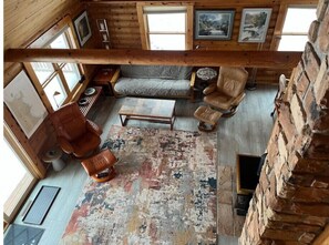 L’intérieur d’un chalet confortable, avec une chambre à coucher située en mezzanine, des murs en bois, une grande fenêtre, un canapé, un fauteuil et une table basse.