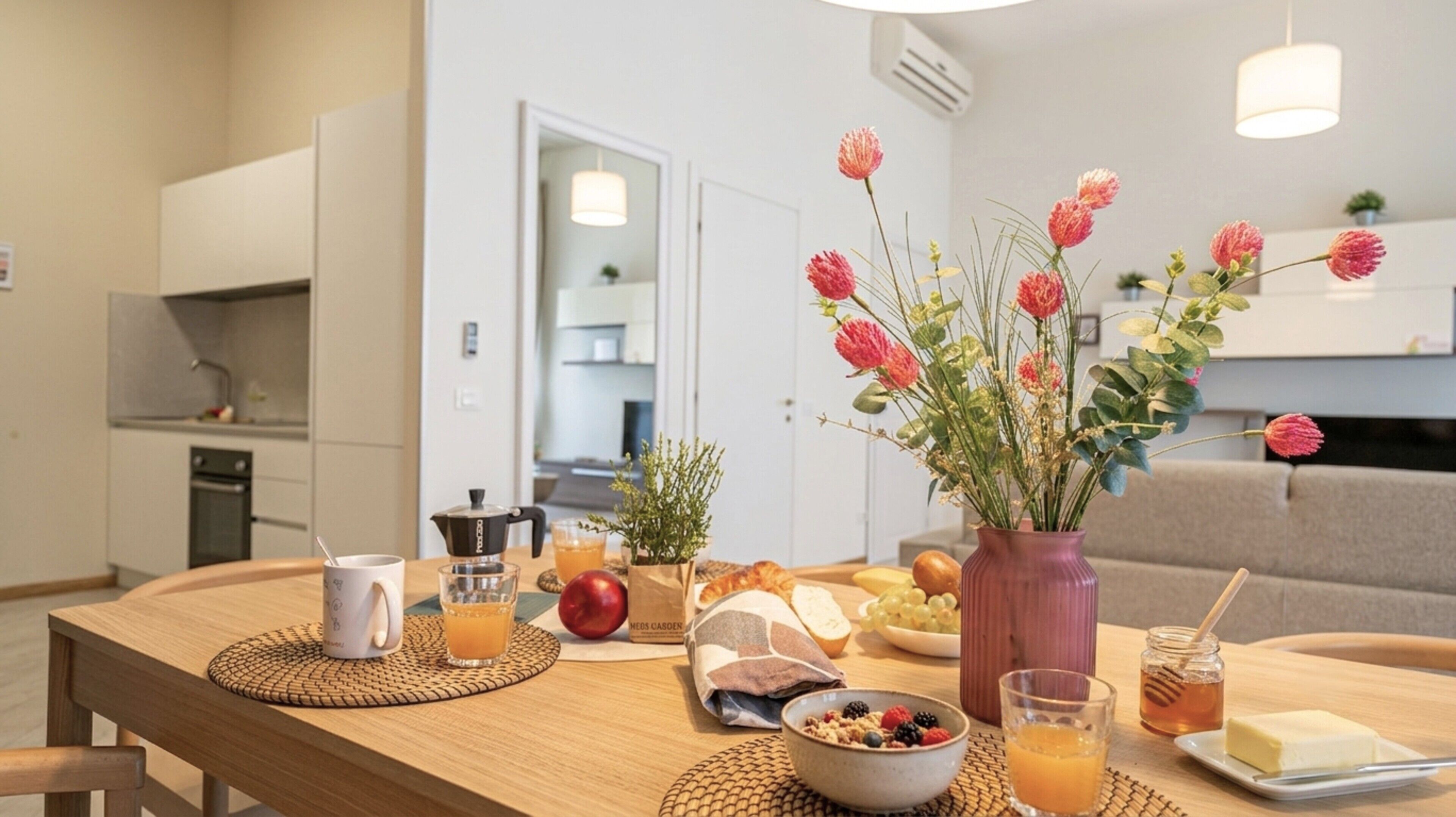 Une table à manger avec des aliments pour le petit-déjeuner, un vase de fleurs et une cuisine en arrière-plan.
