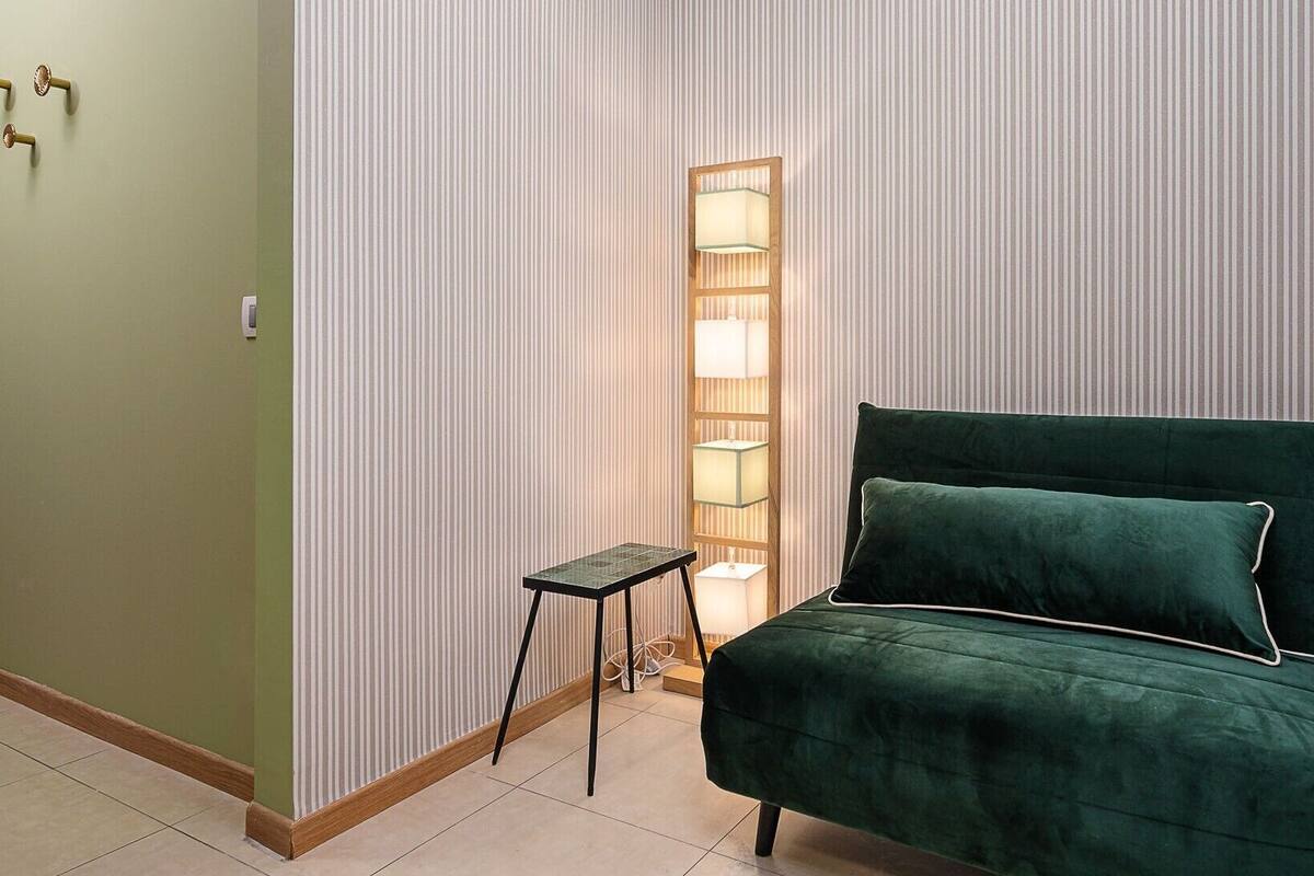 Ein Zimmer mit einem grün gepolsterten Bett, einem kleinen Tisch und einer hohen Stehlampe vor einer gestreiften Tapete.