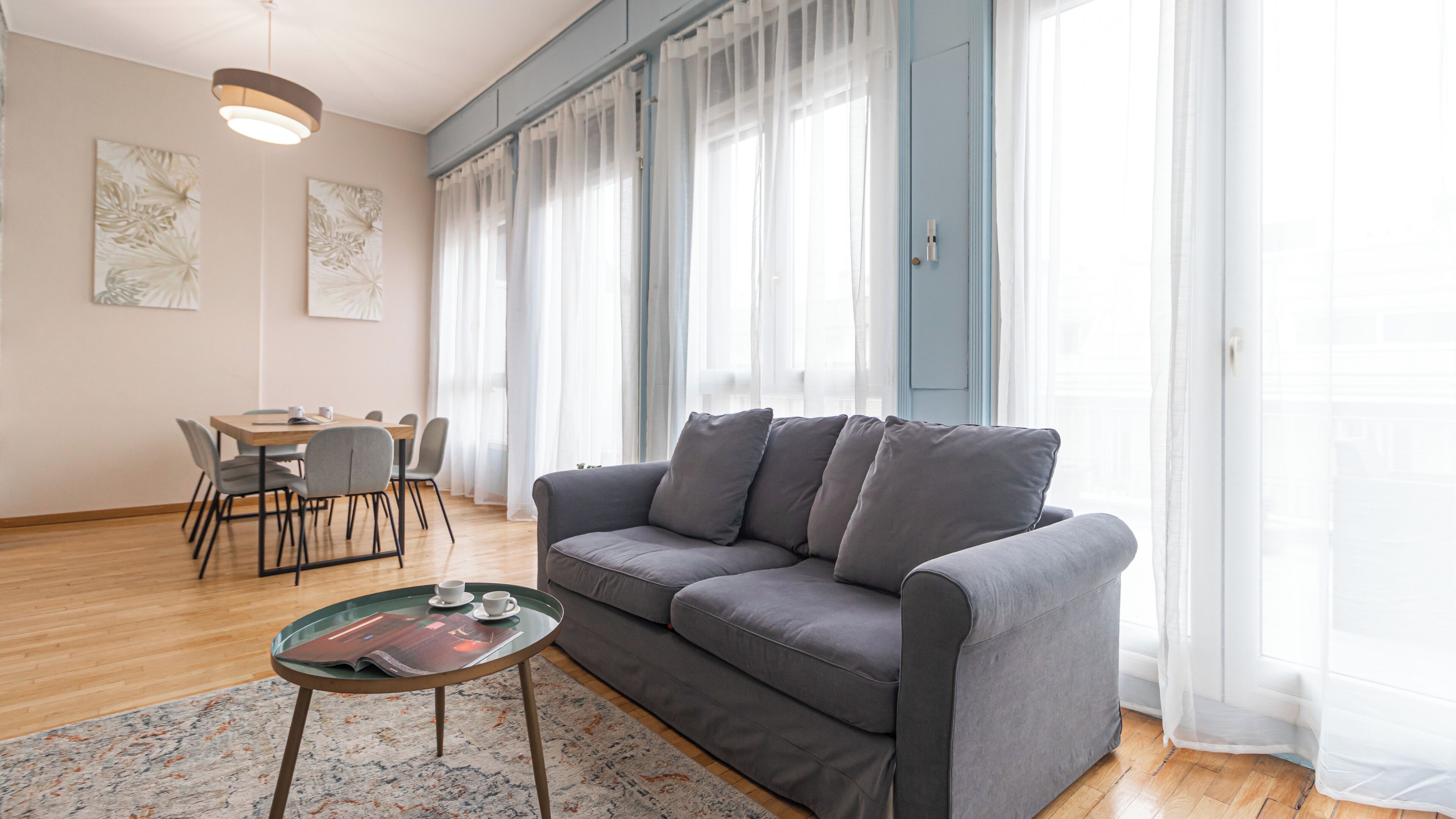 Un salon moderne avec un canapé gris, une table basse en verre, une table à manger avec des chaises et de grandes fenêtres ornées de rideaux légers.