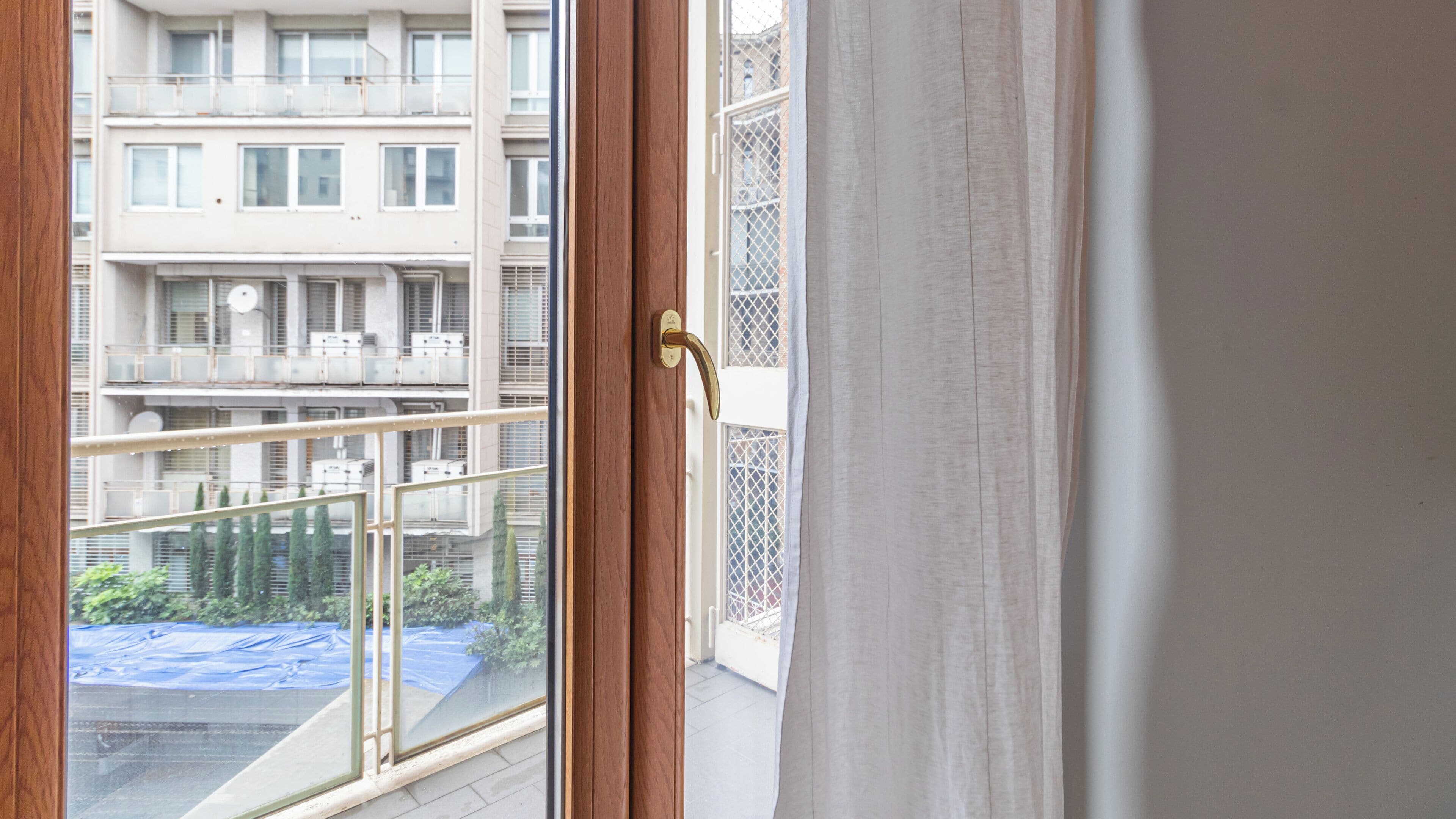 A view through a window with a wooden door and metal railing, overlooking a multi-story residential building with balconies and air conditioning units.