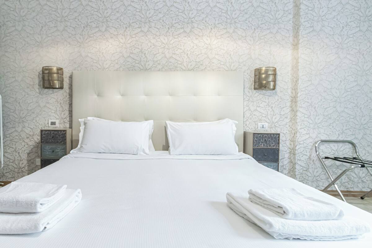 Ein Hotelzimmer mit einem großen Bett, zwei weißen Kissen, einem gepolsterten Kopfteil, zwei Nachttischen mit Lampen, einem grünen Kleiderschrank und gefalteten Handtüchern auf dem Bett.