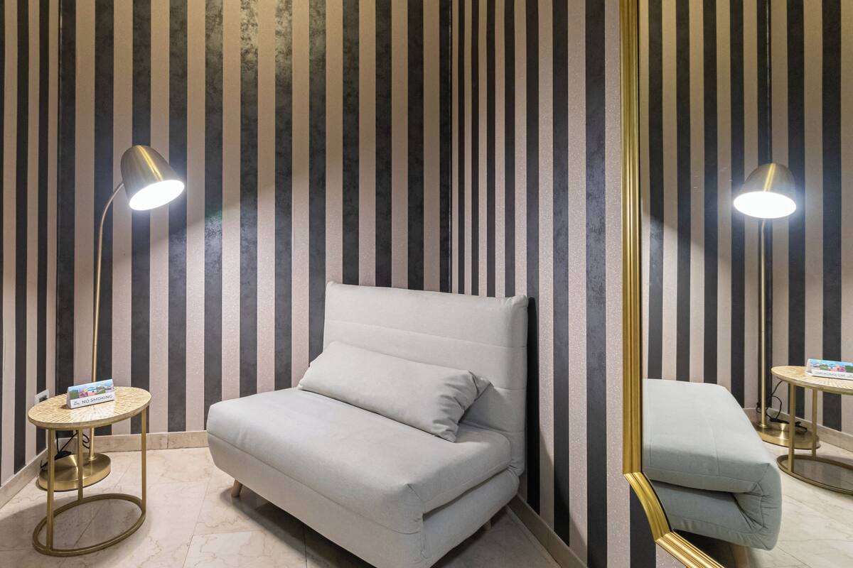 Ein Zimmer mit gestreifter Tapete, einem grauen Sofa, zwei kleinen runden Tischen mit Lampen und einem Spiegel an der Wand.