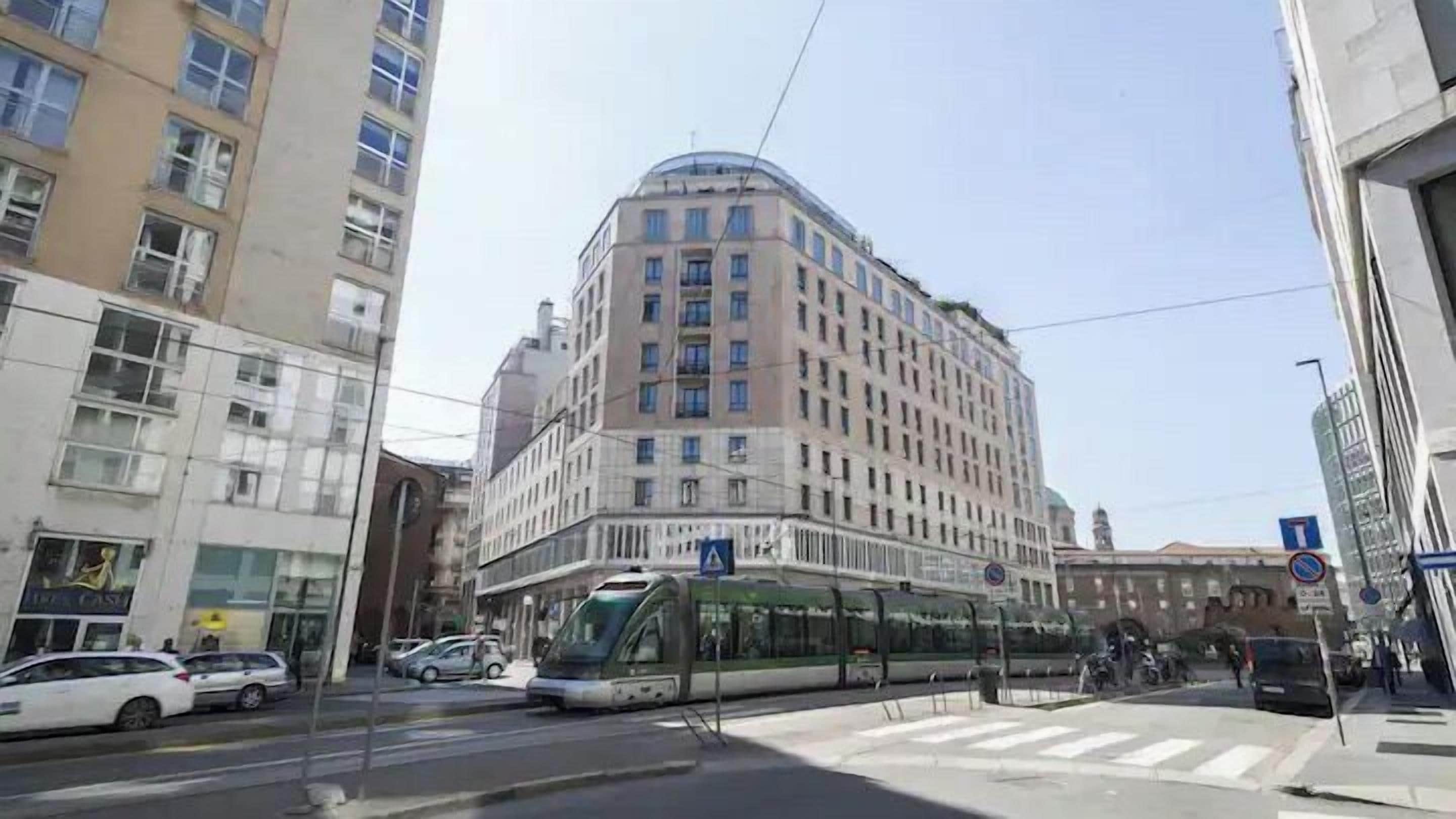 Un bâtiment moderne avec un coin arrondi, un arrêt de tramway et plusieurs voitures garées.