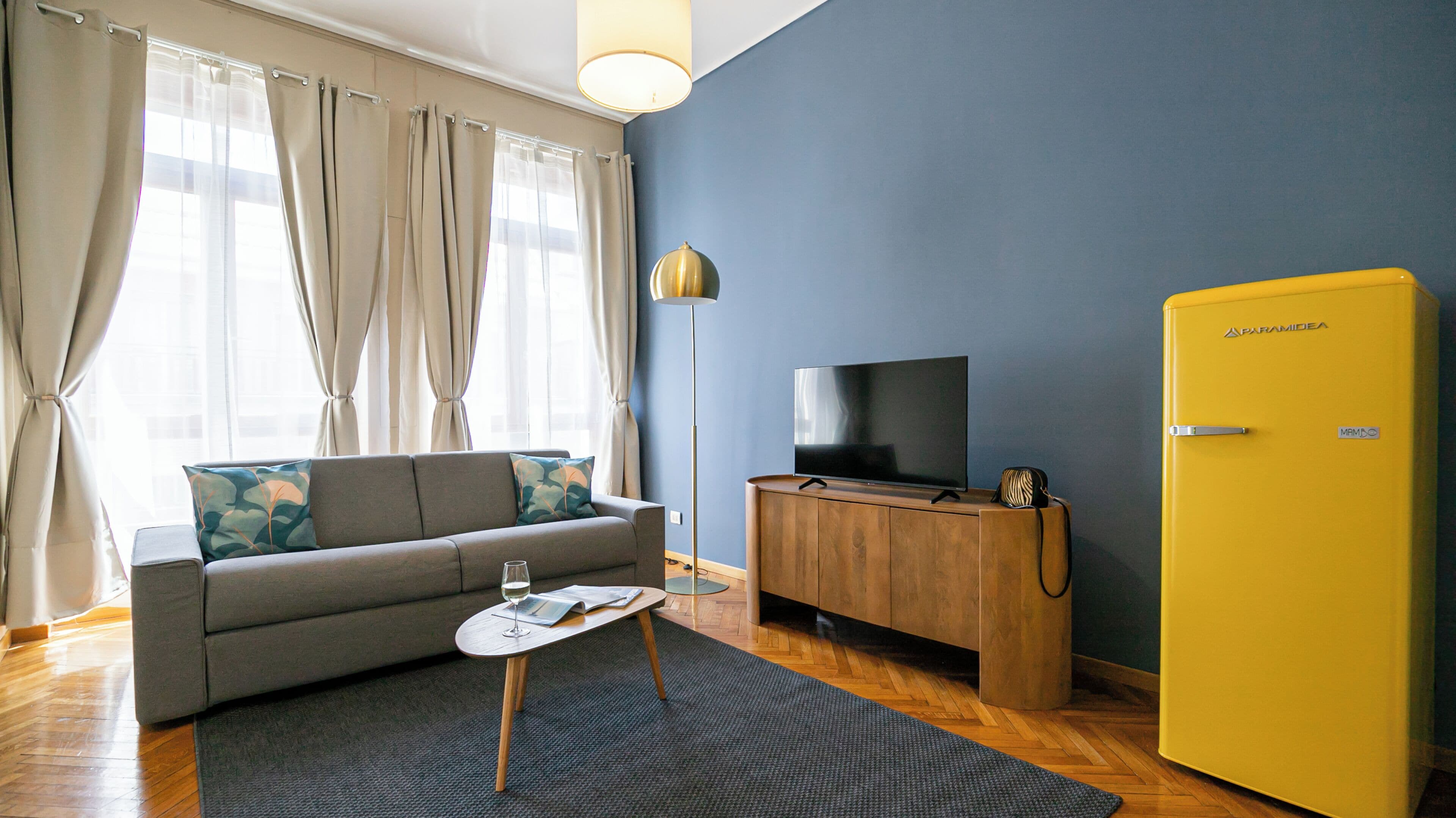 Un salon avec un canapé, une table basse en verre, un téléviseur posé sur un meuble en bois et un réfrigérateur jaune.