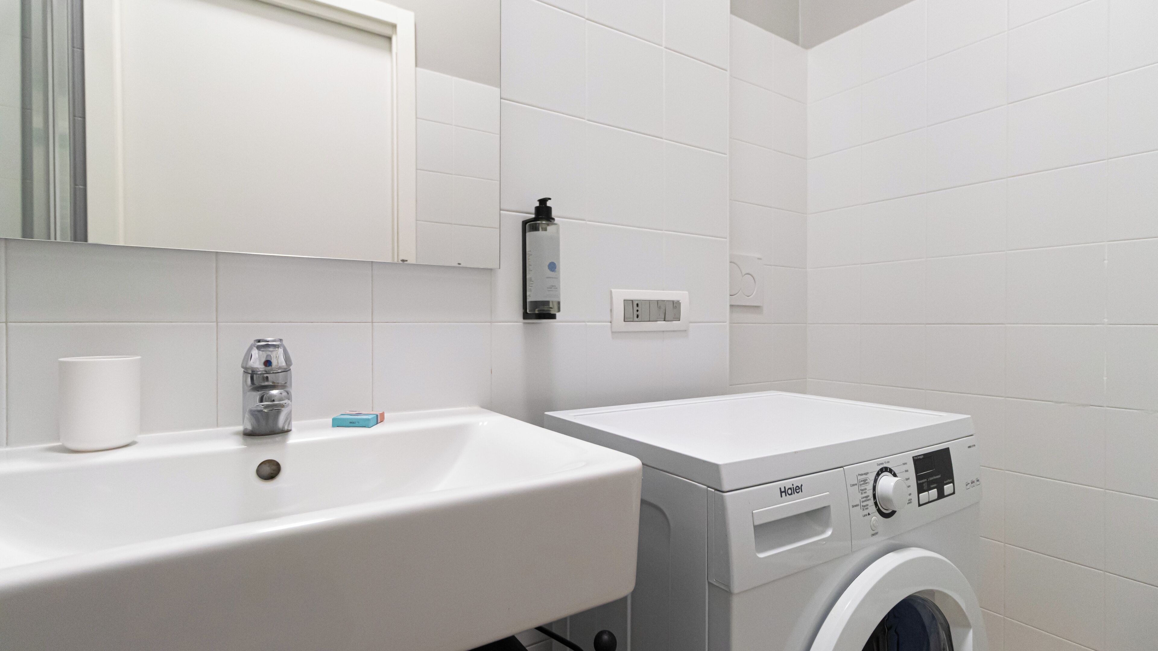 A modern bathroom with a white sink, a mirror, a hand soap dispenser, a soap dish, a white washing machine, and a control panel.