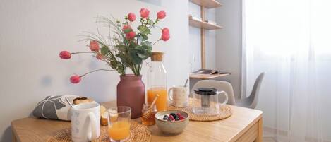 A breakfast setup with a vase of flowers, a bottle of orange juice, a mug, a glass of orange juice, a bowl of berries, and a coffee maker on a wooden table.