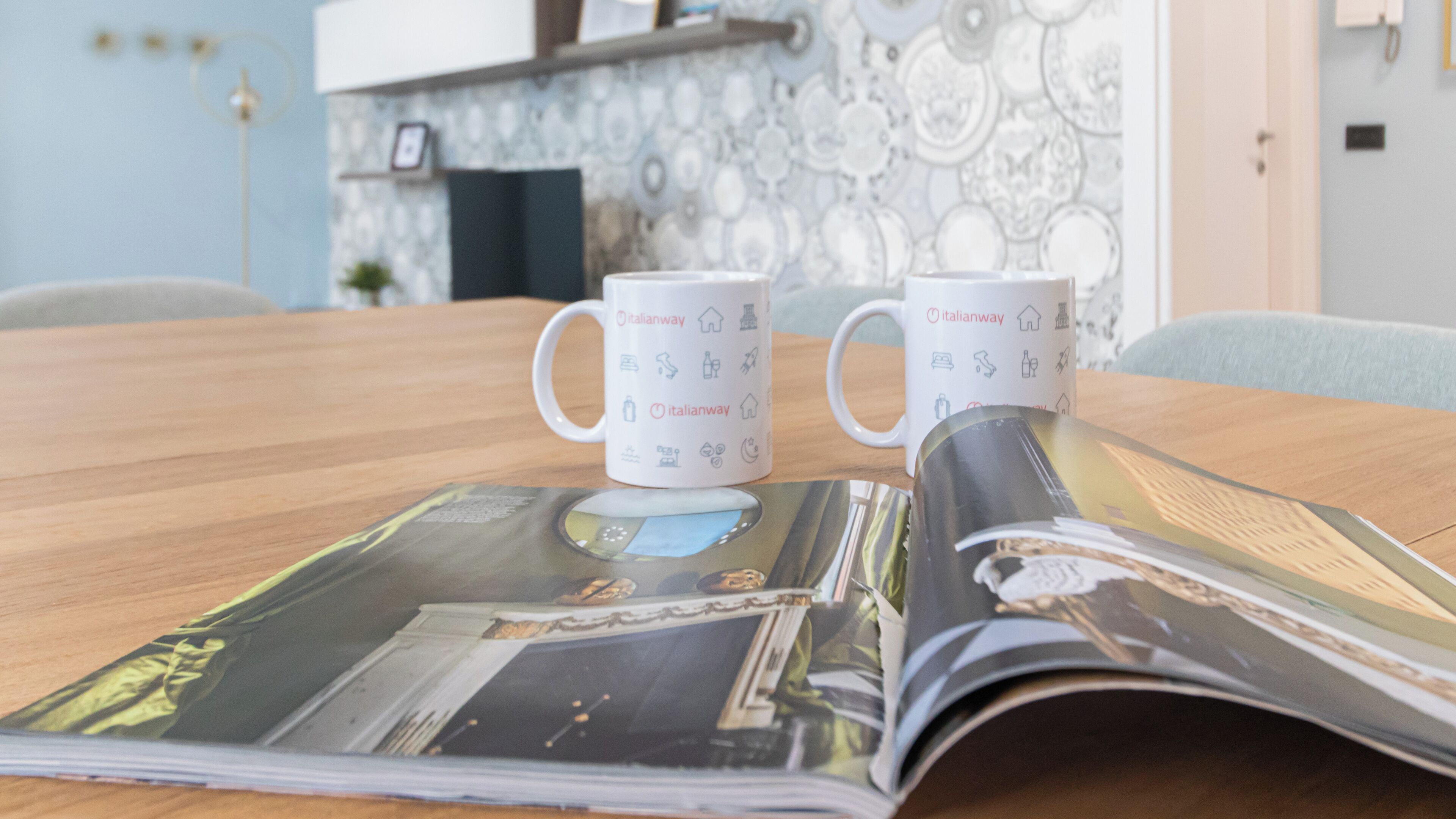Deux tasses blanches ornées du logo Italianway, posées sur une table en bois, à côté d’un magazine.
