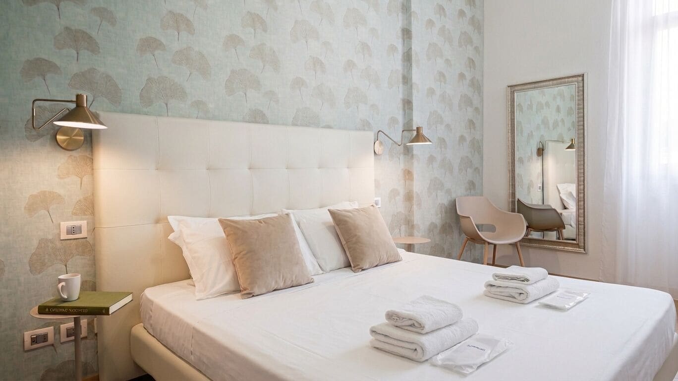 A hotel room with a bed, bedside lamps, a mirror, a chair, and a small table with a book and a cup.