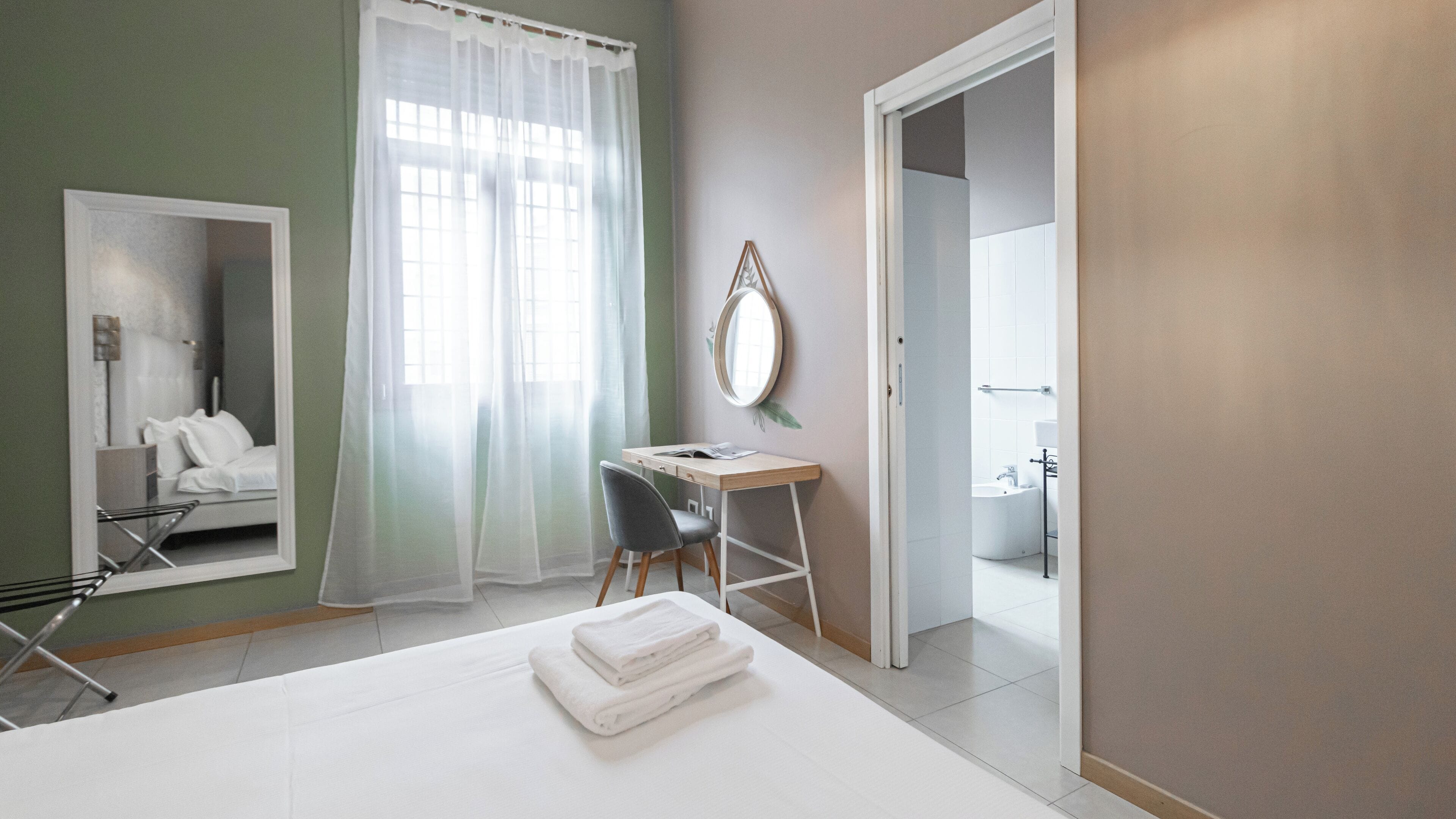 Ett hotellrum med en säng, ett skrivbord, en stol, en spegel och ett badrum som syns genom en öppen dörr.