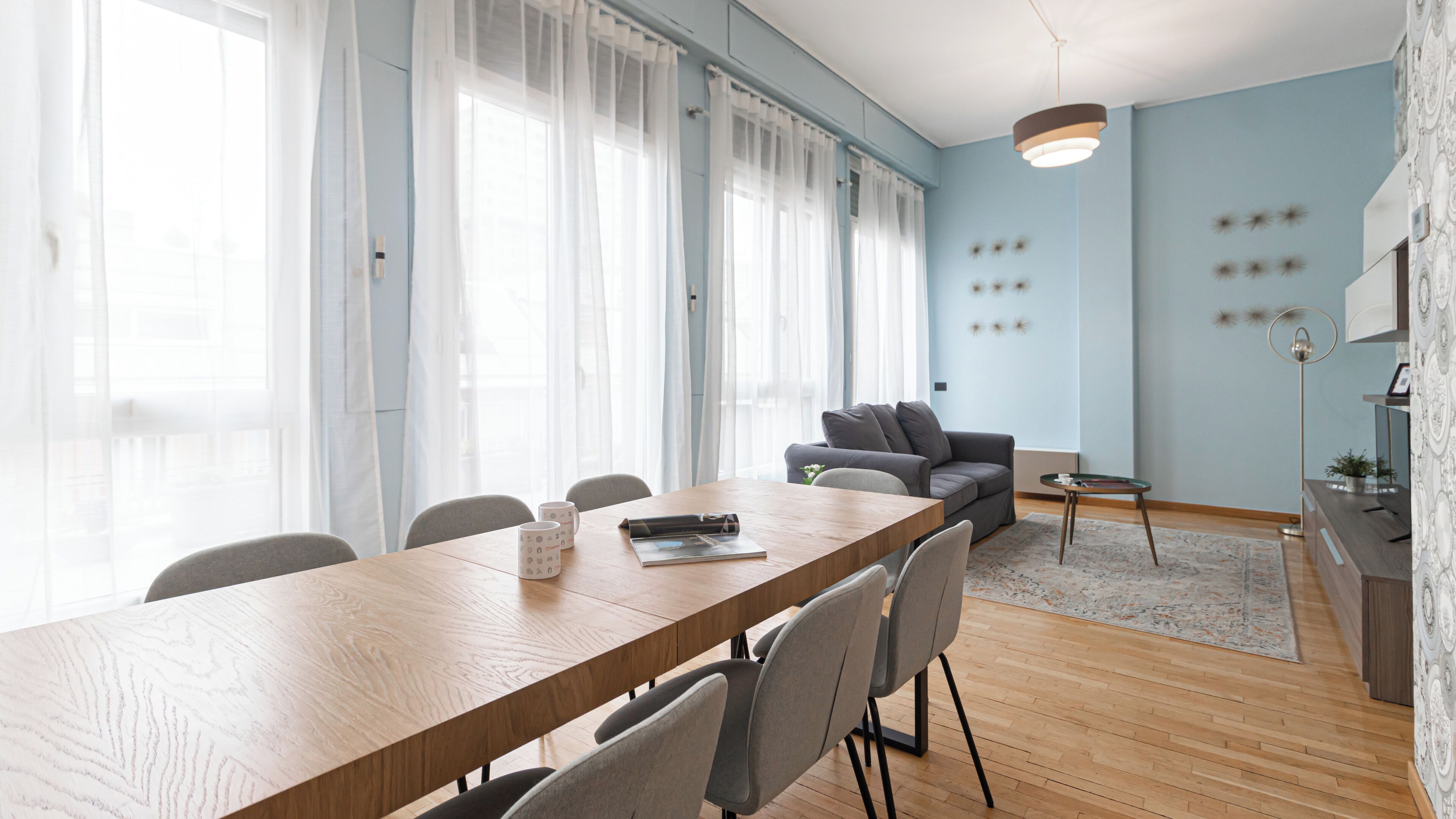 Un salon moderne avec une table à manger en bois, un canapé gris et une grande fenêtre ornée de rideaux légers.