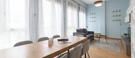 Un salon moderne avec une table à manger en bois, un canapé gris et une grande fenêtre ornée de rideaux légers.