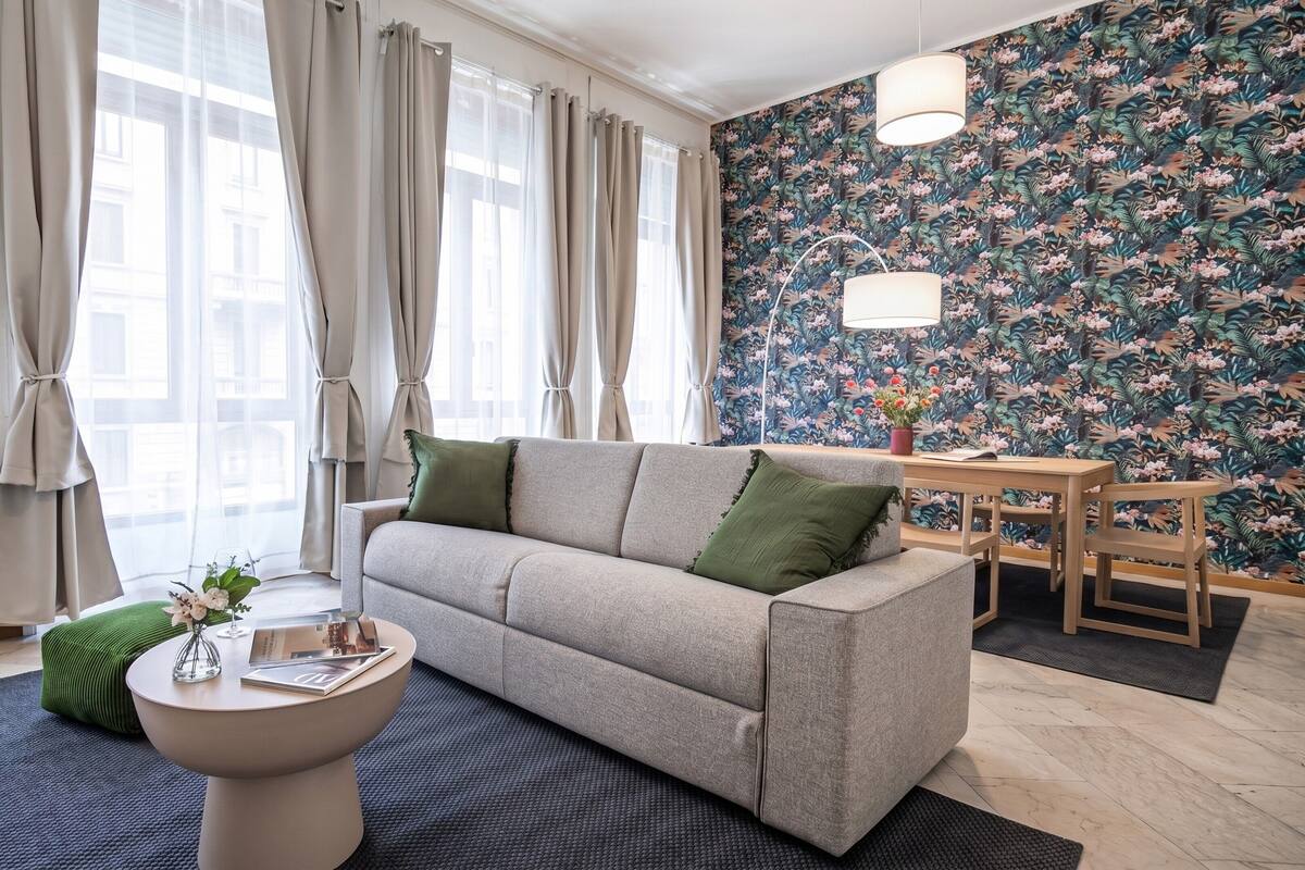 Ein modernes Wohnzimmer mit grauem Sofa, einem runden Couchtisch und floraler Wandgestaltung.