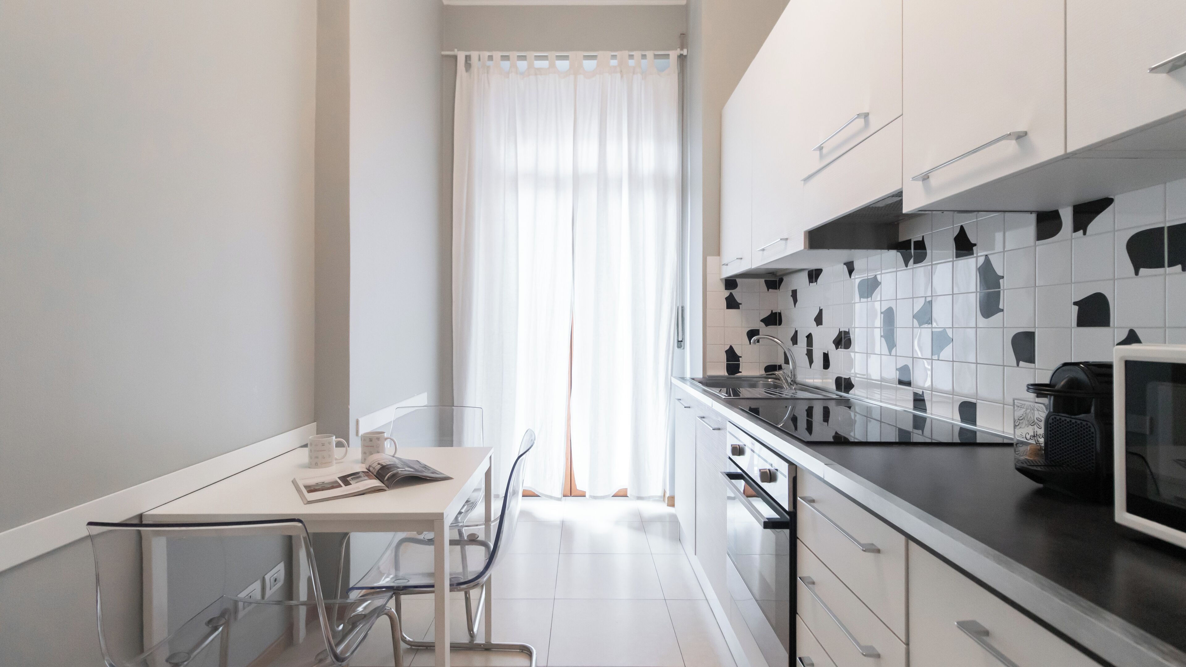 Eine moderne Küche mit weißen Schränken, einer schwarzen Arbeitsplatte und einem Fenster mit durchsichtigen Vorhängen.