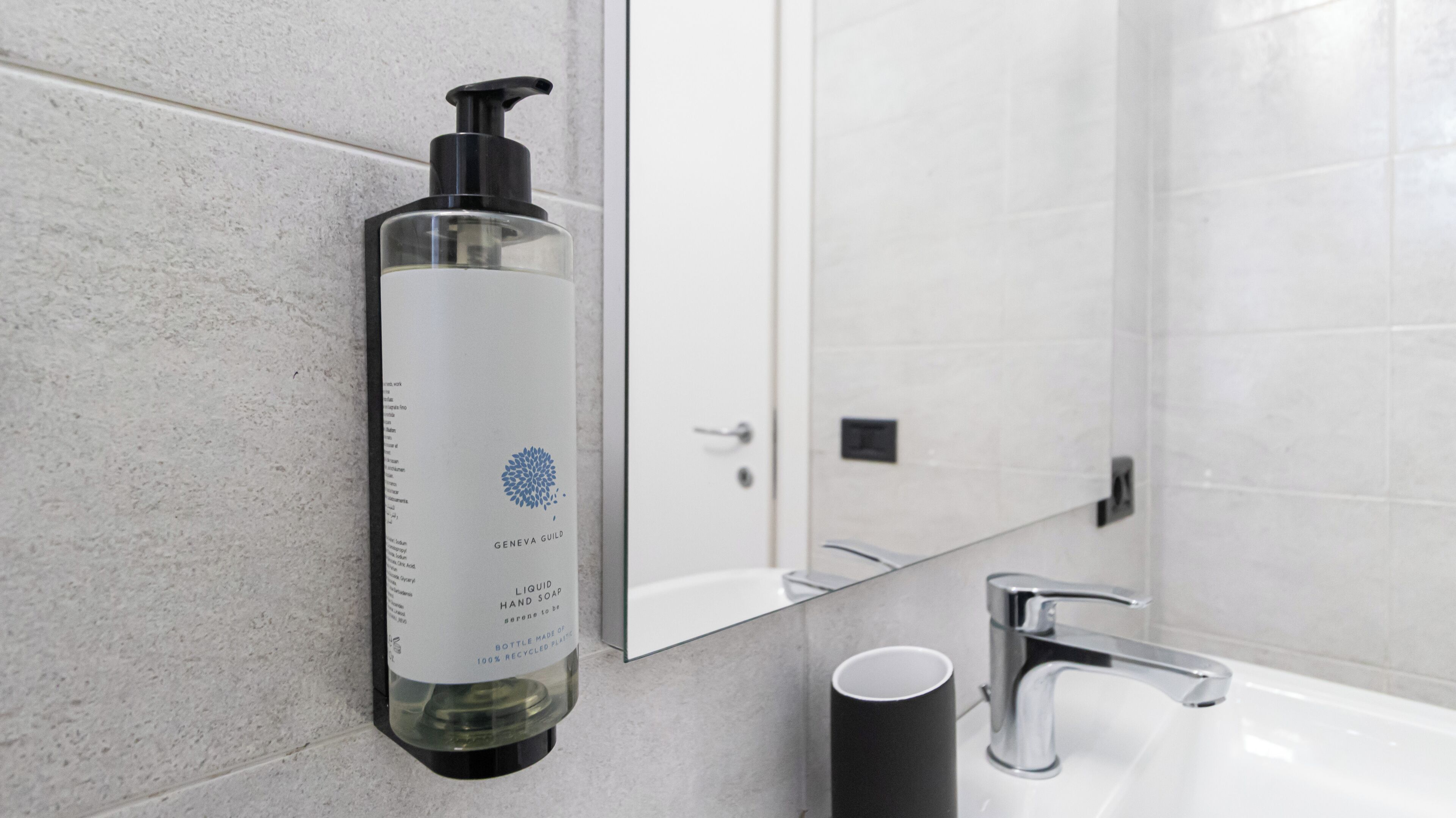 Un baño con dispensador de jabón para manos montado en la pared, un lavamanos y un espejo.