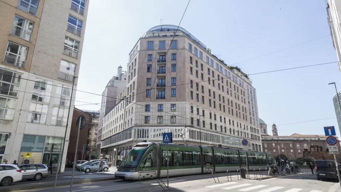 A modern building with a curved corner, large windows, and a tram line running in front.