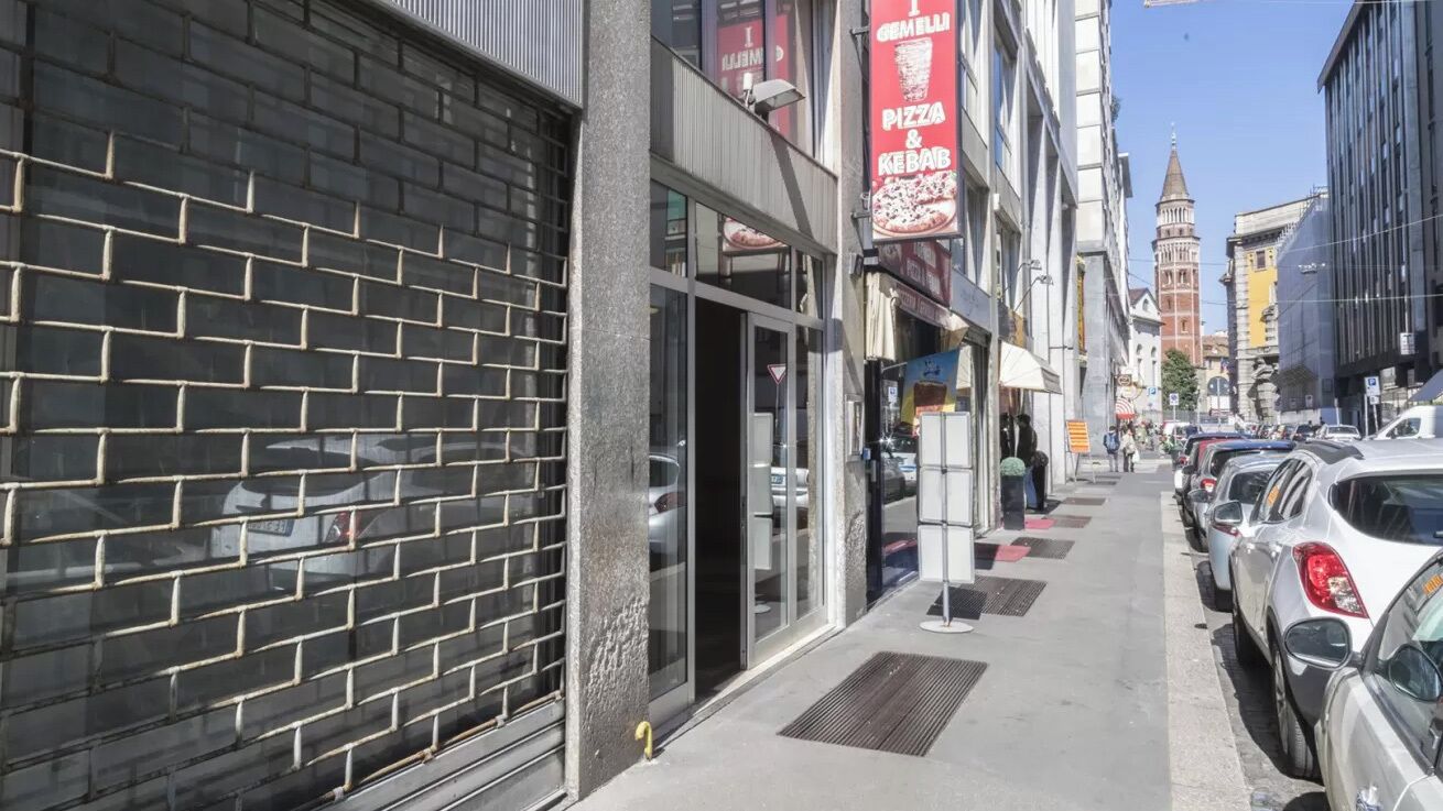 Straatbeeld met een gesloten winkel links, een schoon voetpad, geparkeerde auto's en een historische toren op de achtergrond.