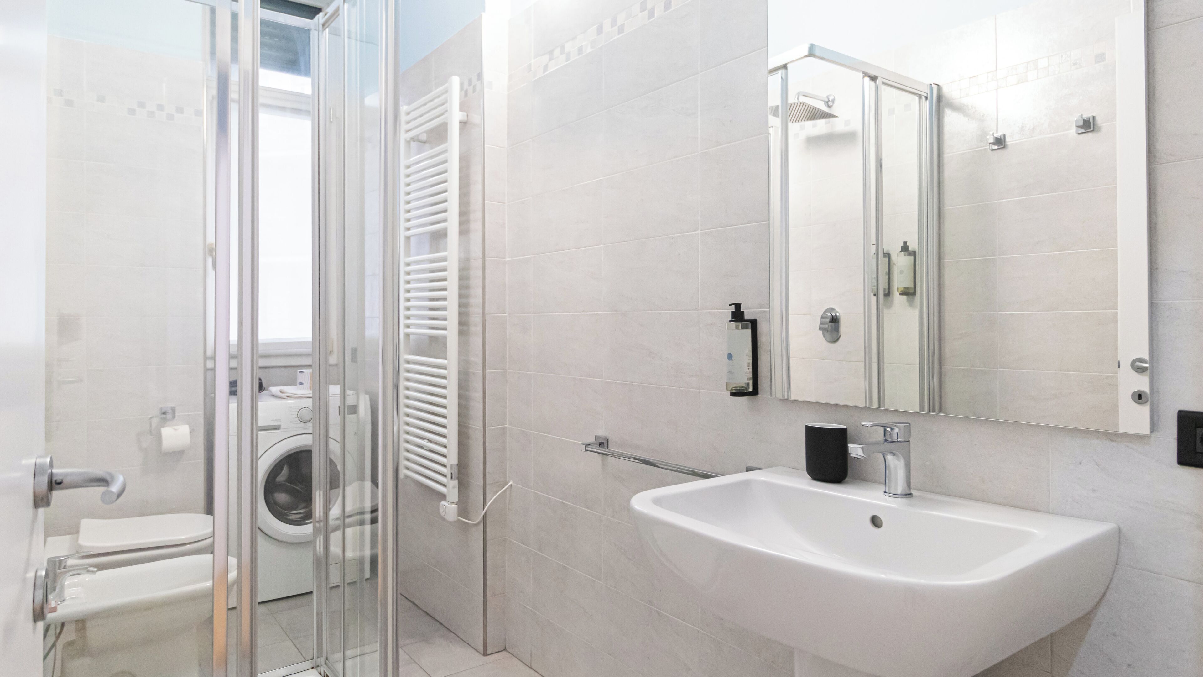 Une salle de bain moderne dotée d’une douche à parois de verre, d’un lave-linge encastré, d’un lavabo blanc et d’un grand miroir.